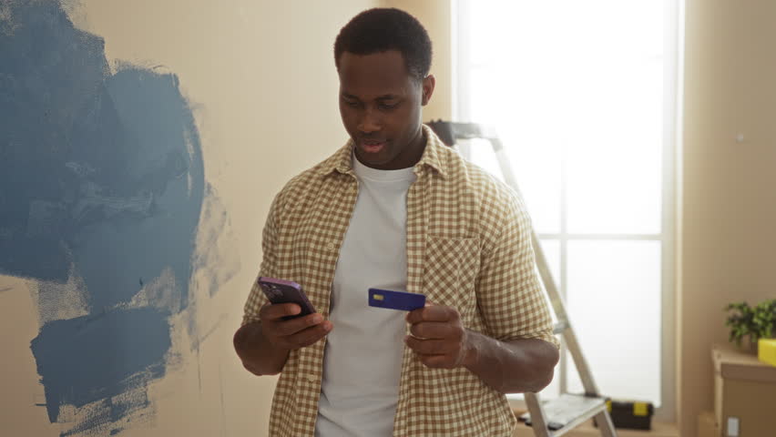 Young man smiling while holding phone and credit card in newly painted living room with ladder in background.