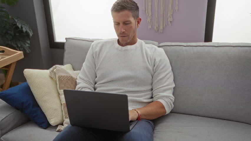 Stressed young man closes laptop on sofa in modern living room, surrounded by cushions, expressing frustration and fatigue.