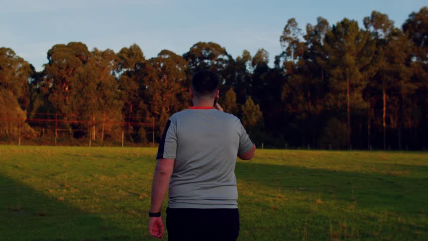 Man walking in grass field, talking on phone, trees and long shadows in background.