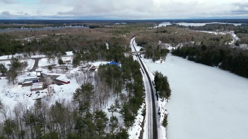 Aerial drone moving in reverse over train railroad tracks and frozen ice Spiers Lake in cottage country Bala, in the Muskoka Lakes region of Ontario, Canada. Evergreen trees and snow during winter.
