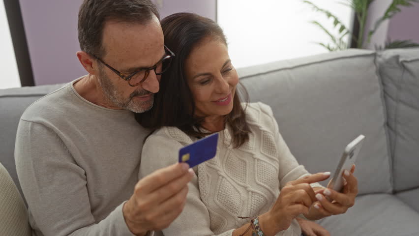 Middle-aged hispanic couple sitting on a sofa in a cozy living room browsing a smartphone and holding a credit card, exuding warmth and family connection.