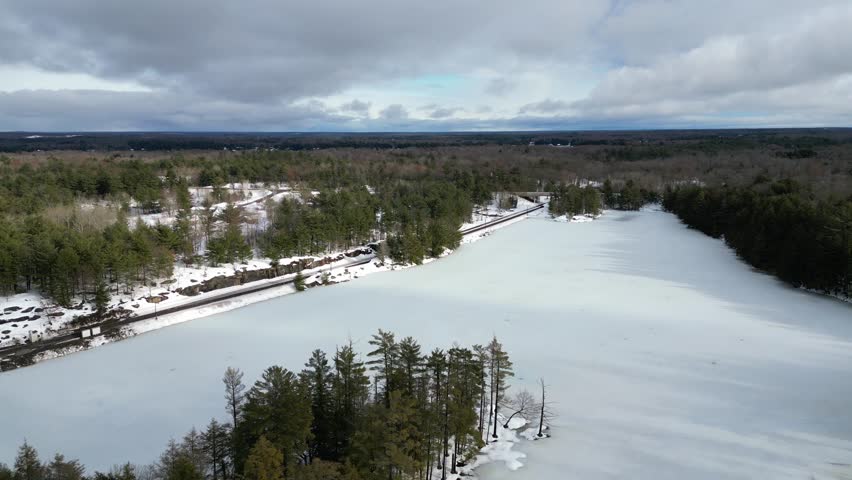 Aerial drone flying over a frozen Spiers Lake towards railroad tracks in rural cottage country Bala, in the Muskoka Lakes region of Ontario, Canada. Evergreen trees and snow during a cold winter day.