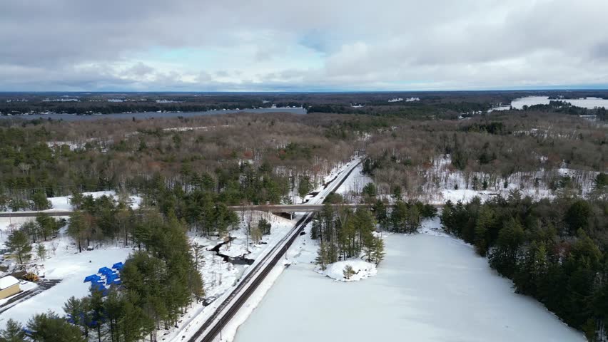 Aerial drone moving in reverse over train railroad tracks and frozen ice Spiers Lake in cottage country Bala, in the Muskoka Lakes region of Ontario, Canada. Evergreen trees and snow during winter.