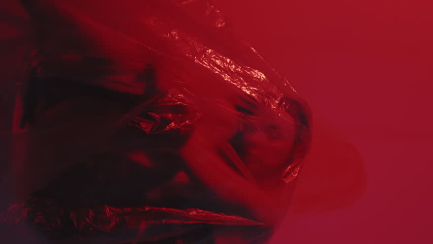 Vertical video. Desperate beauty. Woman panic. Tired girl sleeps sits in polyethylene film dreaming of freedom isolated on dark red pink flicker.