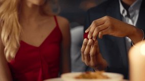 A young, diverse couple enjoys an intimate dinner at a cozy restaurant. The man holds an engagement ring while looking at his partner, who appears surprised and excited, reflecting their love. - Powered by Shutterstock - Get 15% off with code: PIKWIZARD15