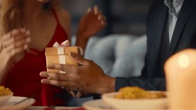 A diverse couple sits closely together at a table in a cozy restaurant, sharing a romantic dinner. The woman hands a beautifully wrapped gift to her partner, filled with affection. - Powered by Shutterstock - Get 15% off with code: PIKWIZARD15