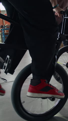 Vertical side tracking slowmo shot of two unrecognizable BMX riders cycling on one wheel of bike, practicing front and backside pivot stunt in warehouse, extreme sports