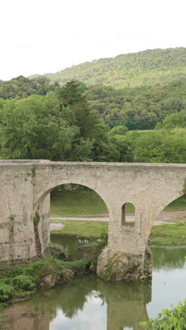 Besalu, Girona, Catalonia, Spain. Famous Landmark Old Medieval Romanesque Besalu Bridge Over The Fluvia River In Cloudy Summer Day.