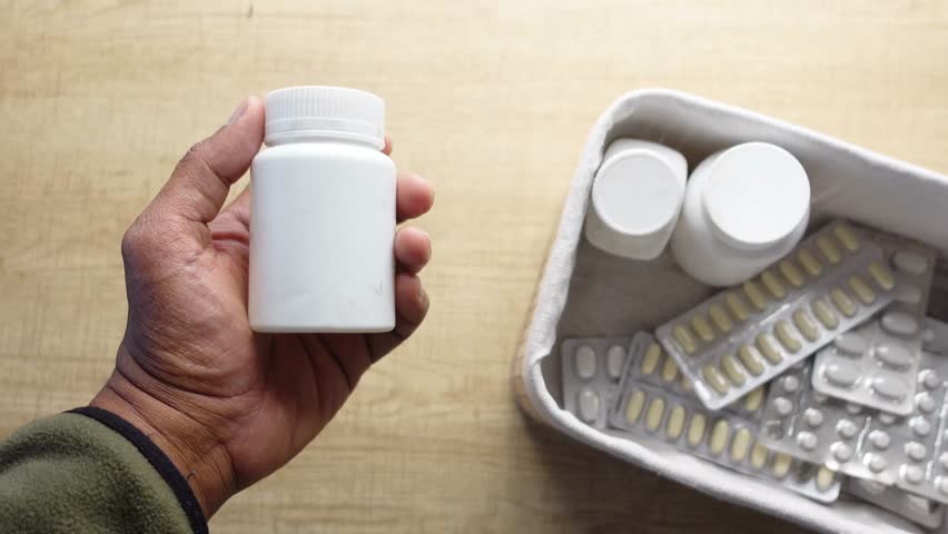 A hand gripping a white pill bottle surrounded by various medications