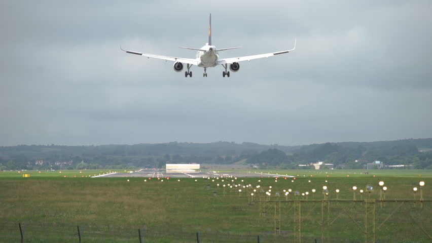 Plane approaches landing on a cloudy day at the airport near a lush green landscape with lights marking the runway