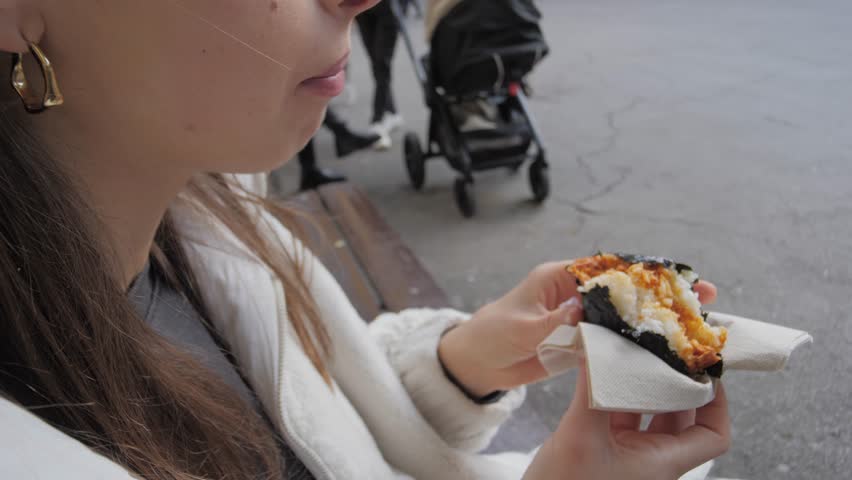 Girl holds and bites eats Onigiri - traditional Japanese Asian snack, close-up. Quick and healthy street food for travelers. Triangular wrap made of nori, rice ball, and delicious varied fillings
