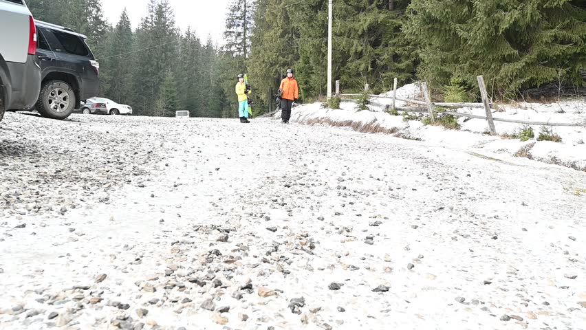 Two skiers walking in a snowy parking lot near a forest, after a day of skiing.