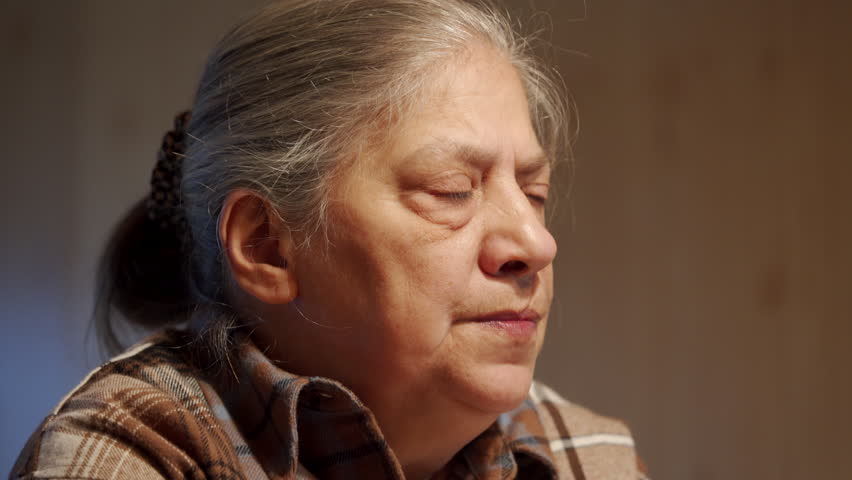 elderly woman sits in a living room and looks at someone with an engaged expression. She appears to be sharing a story, and her eyes seem to hold a wealth of experiences and emotions.