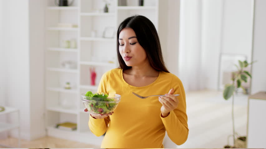 Asian pregnant woman in a yellow top is eating a fresh salad from a bowl while standing in her bright and airy living room. She seems content and focused on her healthy meal choice.
