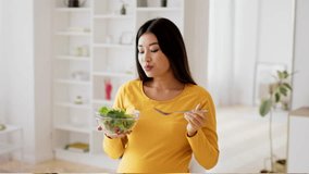 Asian pregnant woman in a yellow top is eating a fresh salad from a bowl while standing in her bright and airy living room. She seems content and focused on her healthy meal choice. - Powered by Shutterstock - Get 15% off with code: PIKWIZARD15