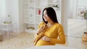 A smiling Asian expectant mother gently holds her sweater while sitting in a bright, modern living room. Natural light fills the space, creating a warm atmosphere. - Powered by Shutterstock - Get 15% off with code: PIKWIZARD15