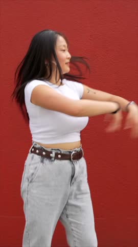 Vertical video of a young asian woman throwing a punch dancing K-pop urban dance outdoors next to red wall