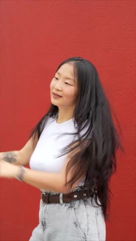 Vertical video of a young asian woman throwing a punch dancing K-pop urban dance outdoors next to red wall