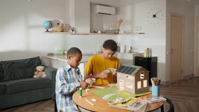 Wide shot of African brothers working together at table, assembling solar-powered model house. Their synchronized efforts reflect strong teamwork and dedication to completing school science project - Powered by Shutterstock - Get 15% off with code: PIKWIZARD15
