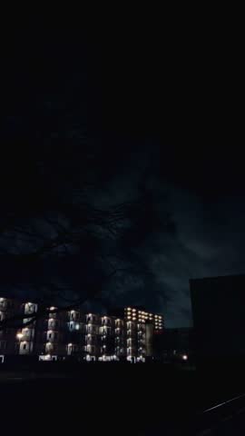 Residential apartment buildings with glowing windows stand under a clouded night sky, contrasting dark tree silhouettes and street lights. Amsterdam, Nederland.