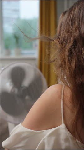 Caucasian woman is suffering from summer heat at home and is sitting in front of fan blown by wind. female housewife holds her hair in her hands for cooling. air conditioning at home