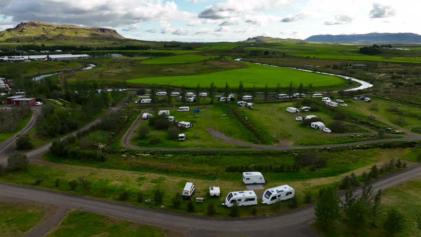 A drone flies over a trailer campsite nestled in green meadows and surrounded by majestic mountains. The stunning scenery captures the peaceful beauty of the camping area.