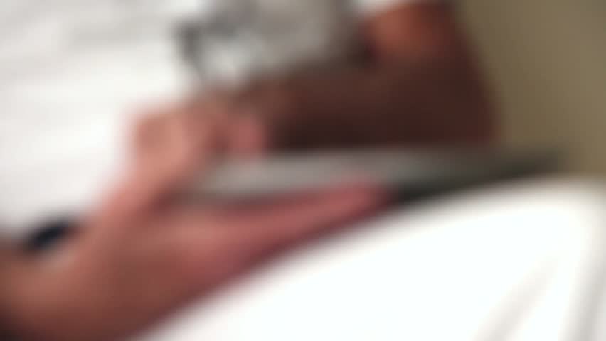 Hands of senior man using tablet sitting on white couch in living room. Starting and ending blurred. (No audio).