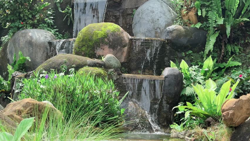 A serene artificial waterfall cascading over moss covered rocks and stone steps,surrounded by lush greenery, tropical plants,and peaceful pond. 