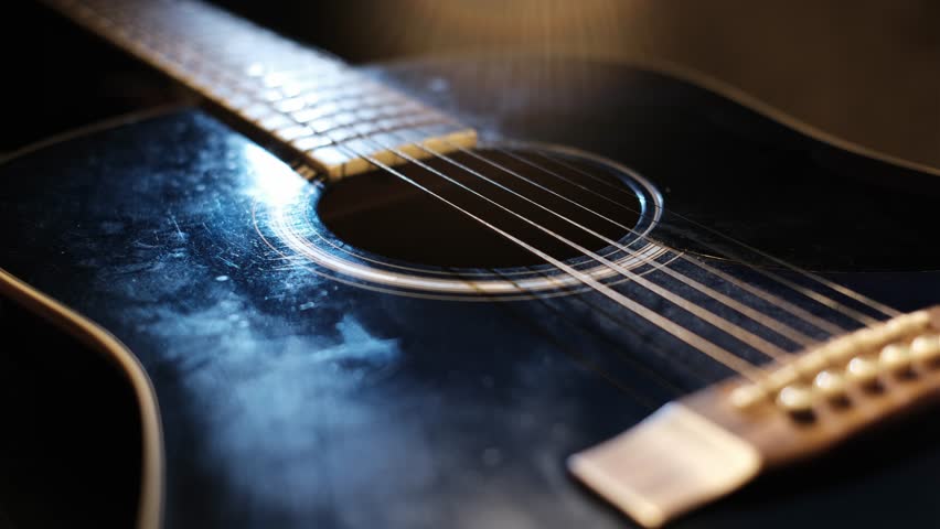 A hand strums a black acoustic guitar, shown in a close-up, capturing the rhythmic motion over the guitar strings, highlighting the textured body and circular opening.