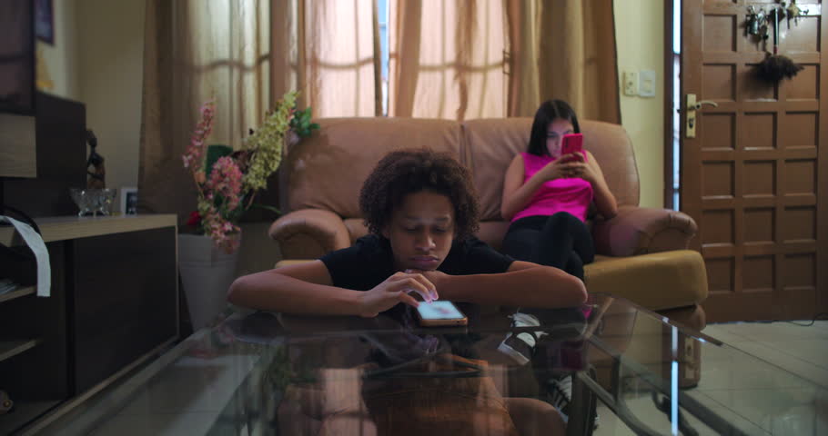 Young boy leaning on glass table staring at smartphone, expressionless, teenage girl behind immersed in her phone, emotional detachment and social media addiction depicted