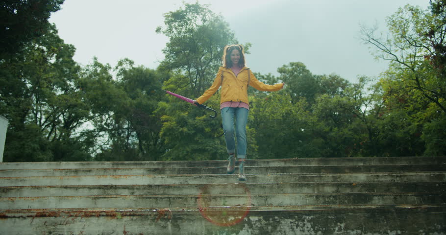 Happy brunette girl in colorful clothes runs up the stairs and jumps on a puddle splashing water in the park after the rain