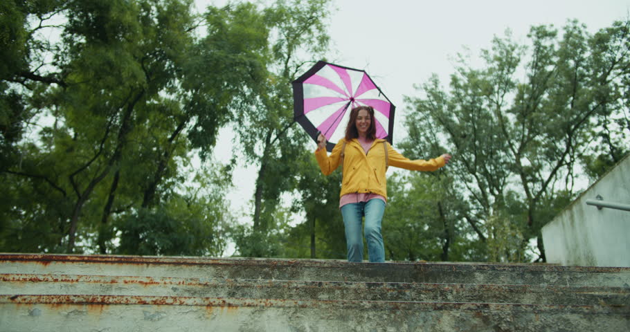 happy brunette girl in multi-colored clothes with an umbrella jumping on a puddle and splashing water in a park with green tall trees in cloudy weather