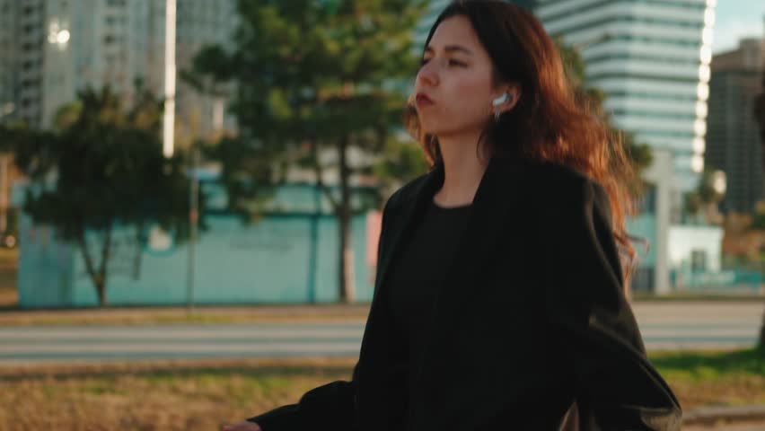 Professional woman wearing wireless earbuds, dancing with joyful energy amid urban park greenery during golden sunset hour, celebrating personal freedom and work-life balance