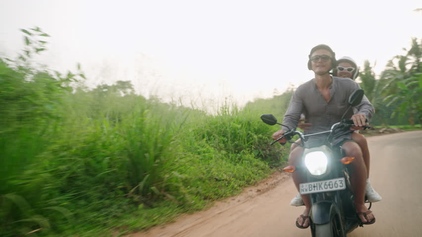 Happy couple ride on motocycle. Lovers travel by motorbike in tropics on sunset. Woman in love spreads hands, laughs in euphoria on jungle road. Nice tourists couple enjoy bike journey. Sun flares.