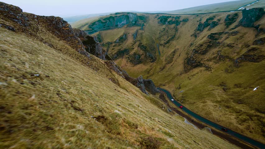 Car driving along winding mountain road surrounded by steep grassy cliffs. Vehicle passing through scenic landscape in Peak District, United Kingdom. Red car traveling on curvy asphalt path through