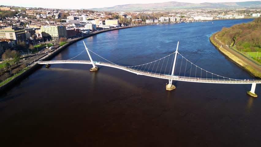 Wide retreating aerial shot of the Peace Bridge in Derry-Londonderry, NI on a bright and sunny day. Filmed in 4K at 30fps and in Rec709 color space.