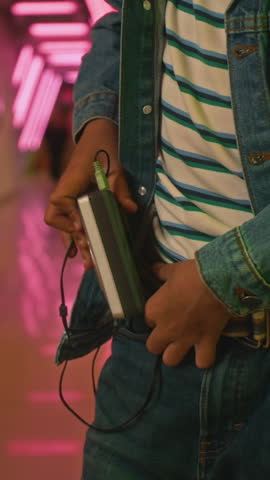 Vertical medium midsection shot of anonymous 1980s teenager in denim jacket and jeans clipping portable audio cassette player to belt, while standing in neon-lit corridor indoor