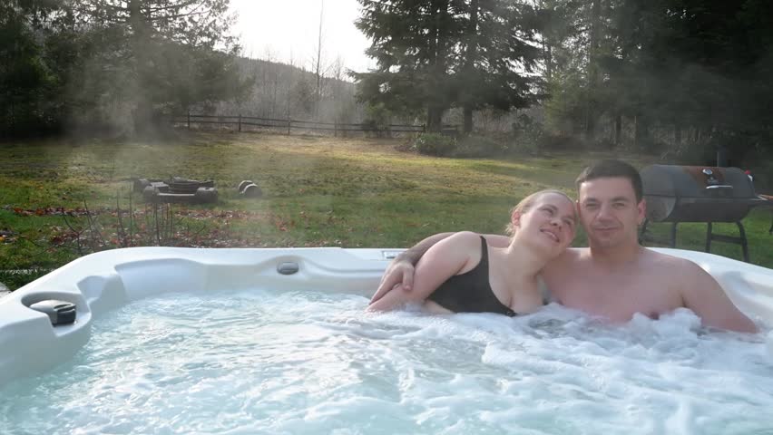 A young couple of a man and a woman are relaxing in a jacuzzi on a weekend, gently embracing. The background is a beautiful landscape of nature.