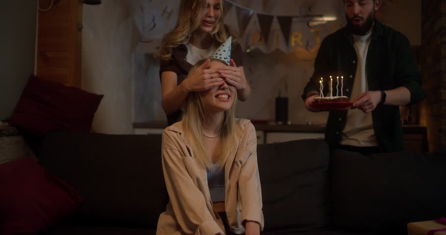 Happy blonde girl in a party cap receives a surprise from her friends and blows out the candles on the cake during a party at home