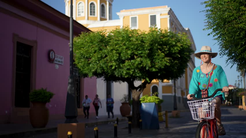 A mature woman experiences the vibrant streets of Granada, Nicaragua while biking. The colorful buildings and lush greenery create a lively atmosphere.