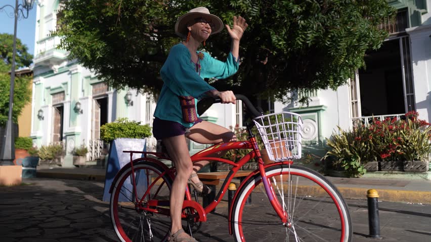 A mature woman joyfully rides a bright red bicycle along the cobblestone Calzada street of Granada, Nicaragua, soaking in the local culture and scenery.