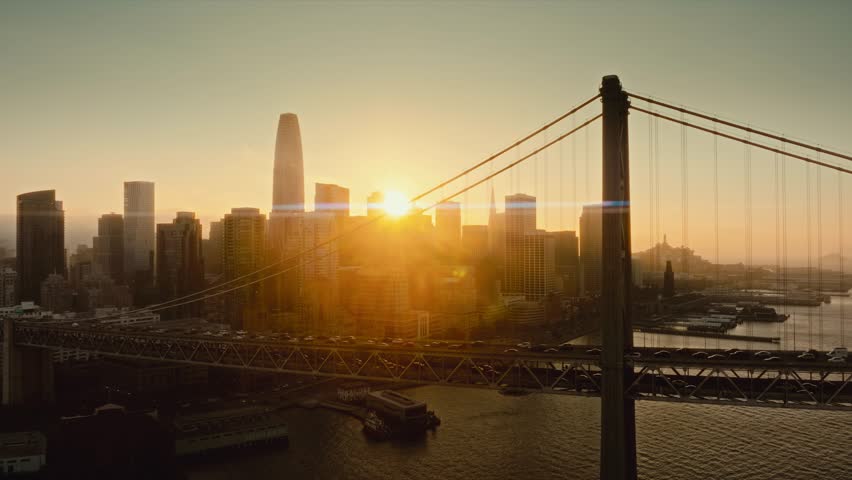 4K Drone Aerial footage flying over the Bay Bridge, San Fransisco at sunset. Sun setting, flaring between the bridge trestles. Bay Bridge in foreground, Downtown San Francisco just behind it.