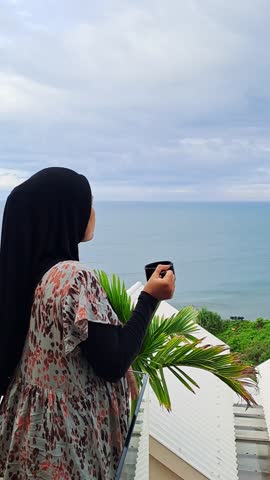 A woman in a hijab enjoys a peaceful morning, sipping from a cup while overlooking a serene ocean view. The tranquil scene captures a moment of quiet contemplation and natural beauty.
