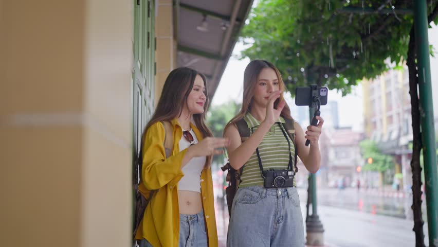 Two Asian women using phone record vlog while traveling during raining. Attractive tourists feel happy and relax while walking along the street, enjoying leisure time during holiday vacation together.