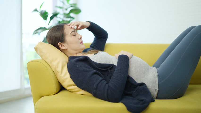 Asian pregnant woman lying on sofa and holding her head with pained expression. woman experiencing headache or dizziness due to pregnancy, hormonal, fatigue, rest and medical attention at home.