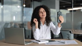 Curly haired businesswoman talks on a video call in the office. She uses her smartphone and sits at the office desk. - Powered by Shutterstock - Get 15% off with code: PIKWIZARD15
