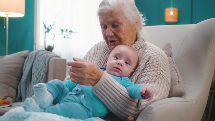 An 80-year-old great-grandmother is sitting in an armchair and holding her great-grandson's 3-month-old newborn baby in her lap at home