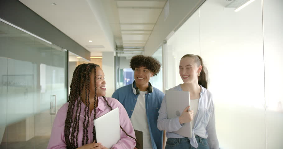 Walking in hallway, three students carrying laptops and tablets, smiling and talking. Education, technology, school, friendship, learning, digital