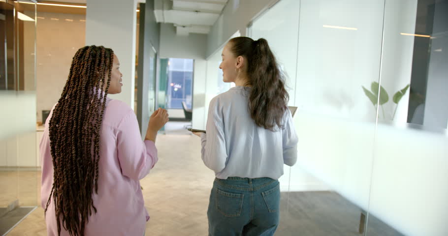 Walking in office hallway, two women discussing work and holding smartphone. Business, colleagues, communication, corporate, teamwork, technology