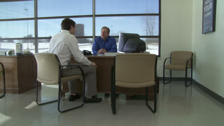 Customer discusses business with bank employee at a desk. Camera moves to follow